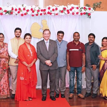 Group photo of eight people standing on a red carpet with floral decorations behind.