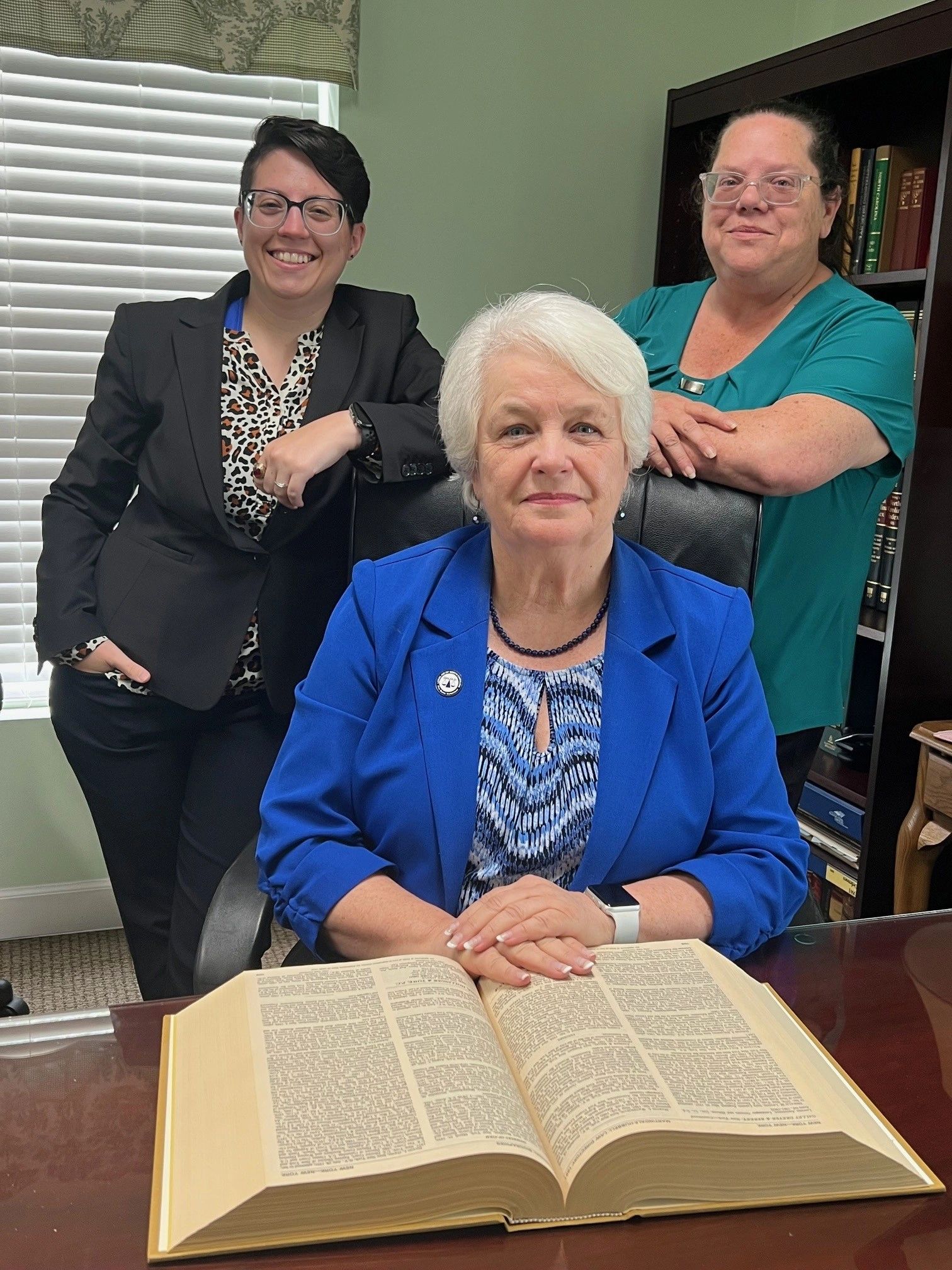 Three women in an office pose with an open large book on the desk.