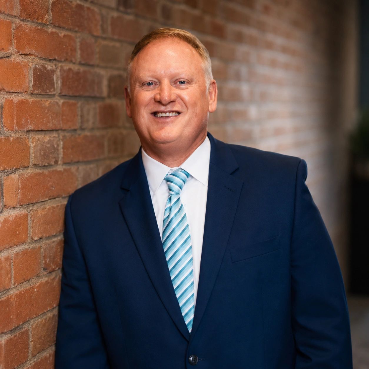 Man in blue suit smiling against a brick wall.