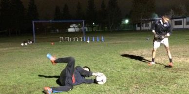 Two people practicing soccer goalkeeping on a field at night.