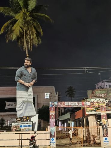Night view of a large statue of a man in traditional attire near a palm tree and entrance gate.