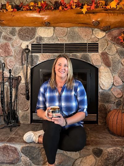 Woman in blue plaid shirt with cup of coffee sitting in front of a fireplace