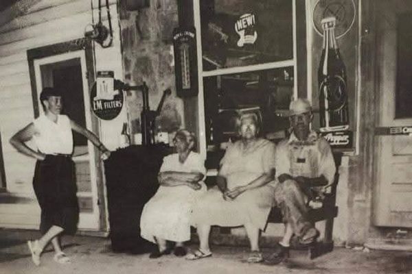Four people in vintage attire pose outside a store, three seated and one standing.