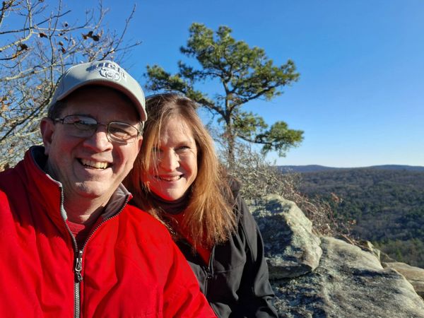 A smiling couple takes a selfie on a sunny rocky overlook with a forested landscape behind them.