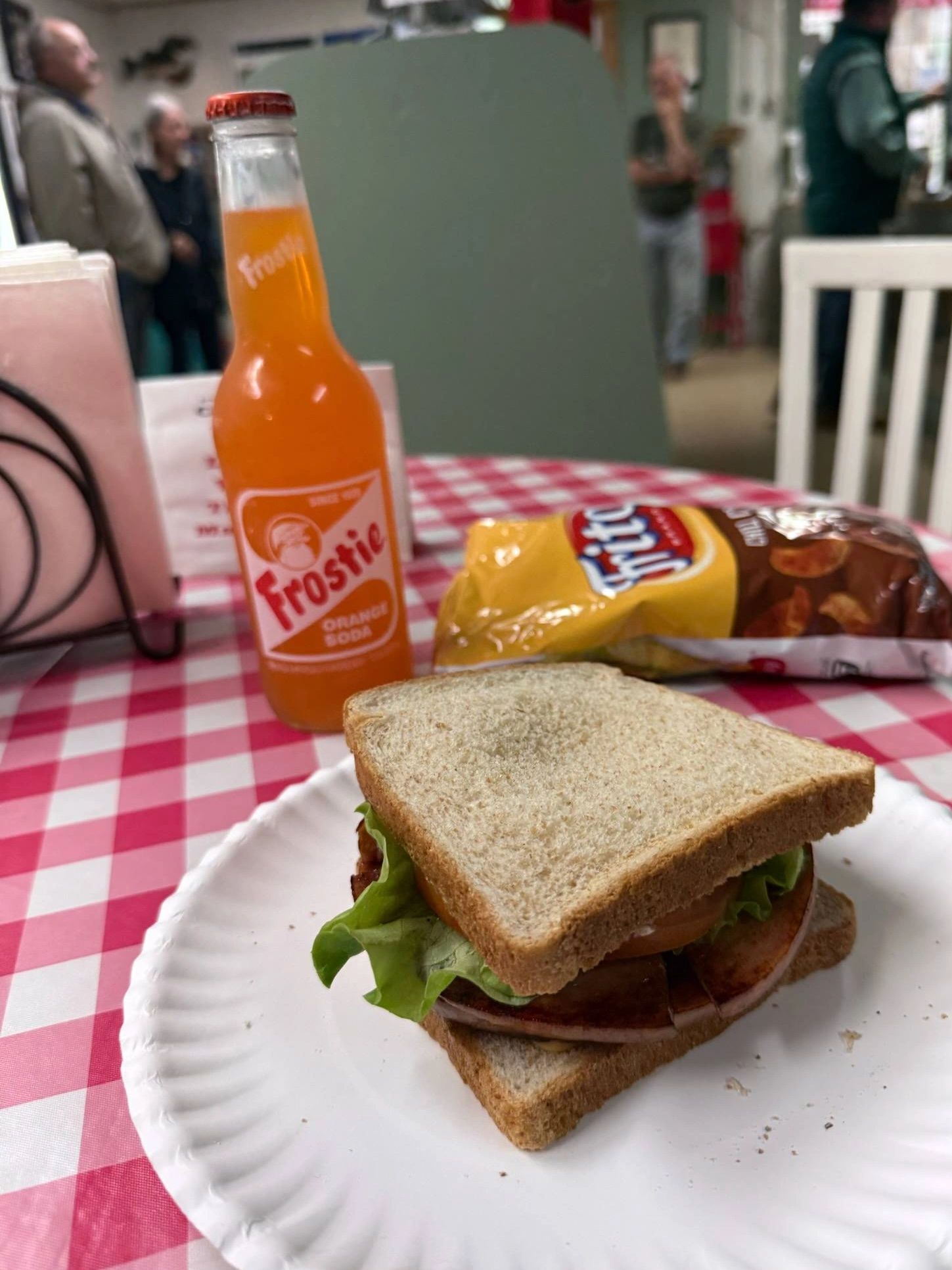 A sandwich with lettuce and tomato on wheat bread, an orange soda, and a bag of chips on a checkered table.