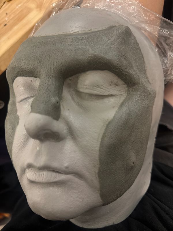 A white face mold with gray textured clay applied on forehead, eyes, and cheeks.