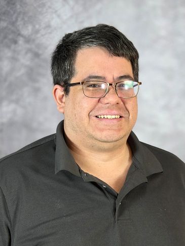 A man with glasses smiling, wearing a dark polo shirt against a gray backdrop.