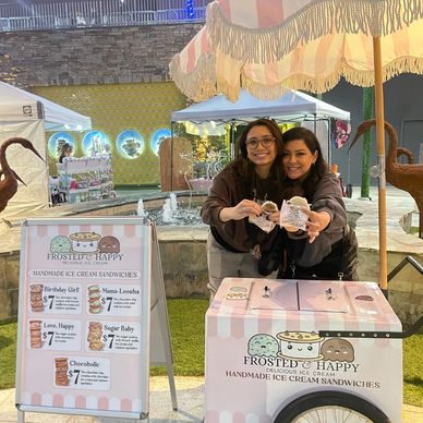 two girls eating ice cream sandwiches and holding sandwiches behind ice cream cart