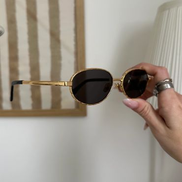 Hand holding gold-framed sunglasses with dark lenses indoors.