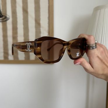 Hand holding brown Chanel sunglasses with logo on the side.