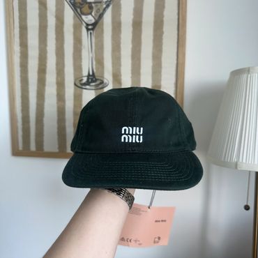 Black Miu Miu cap held indoors against a striped artwork background.