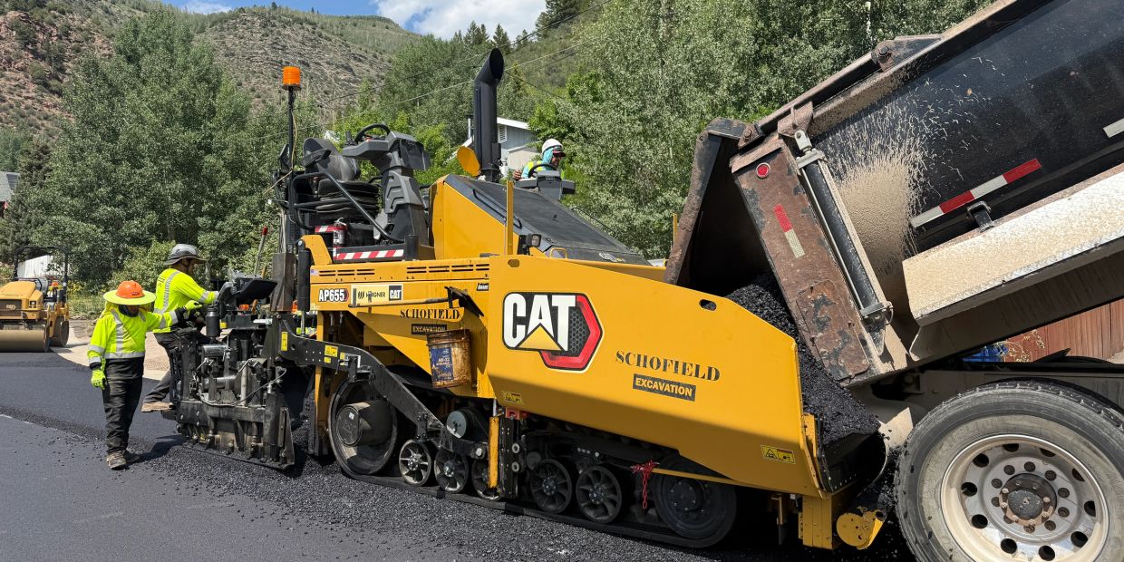 Workers using a yellow CAT machine to pave asphalt on a road in a mountainous area.