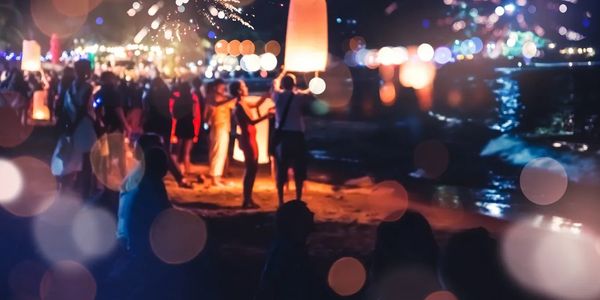 People releasing glowing lanterns on a beach at night with fireworks in the background.