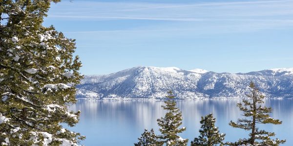 Snow-covered trees frame a clear lake with snowy mountains in the background.
