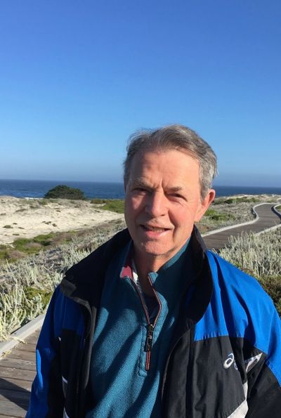Man in a blue jacket standing on a boardwalk near the ocean under a clear sky.