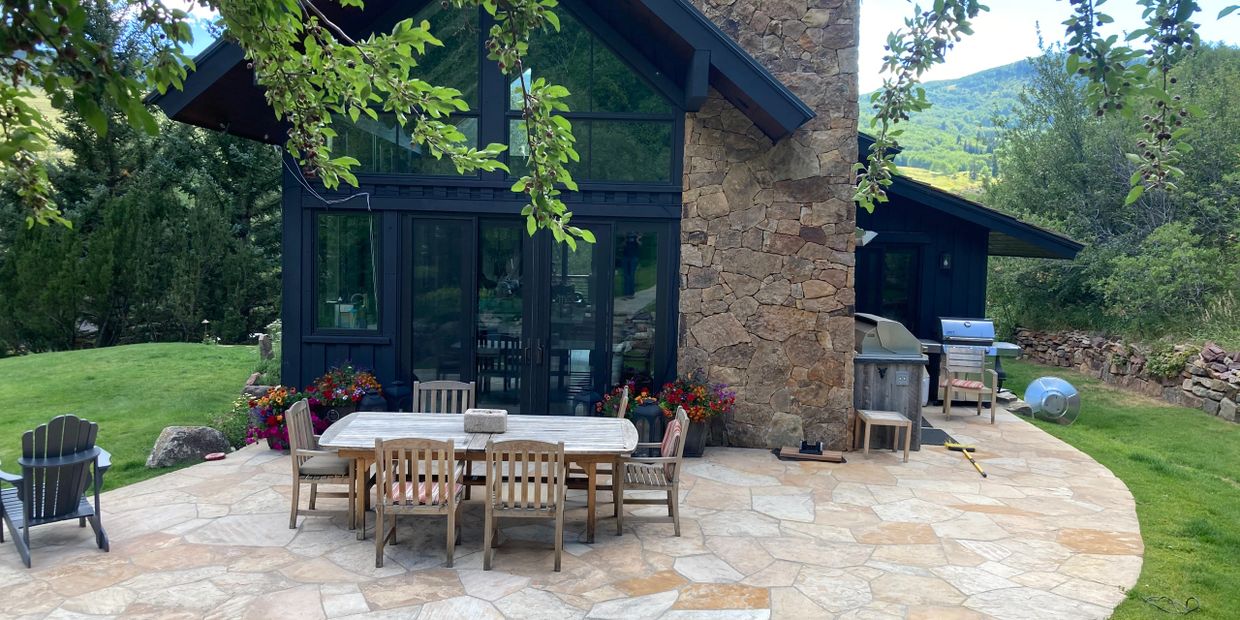 Outdoor patio with wooden table, chairs, and stone fireplace in a lush green mountain setting.