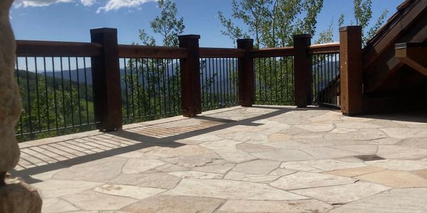 Sunlit stone patio with wooden railing overlooking green hills and blue sky.