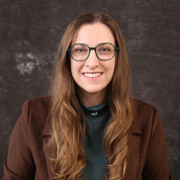Smiling woman with long hair and glasses wearing a brown jacket.
