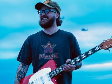 Man with tattoos smiling while holding a red electric guitar outdoors.