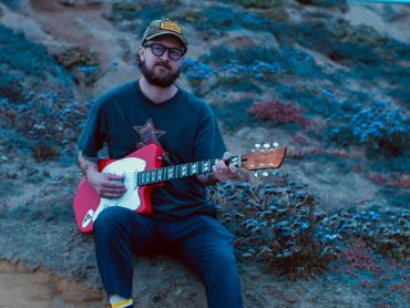 Man playing red electric guitar outdoors in a rocky area.