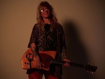 Woman with tattoos holding an electric guitar against a dark background.