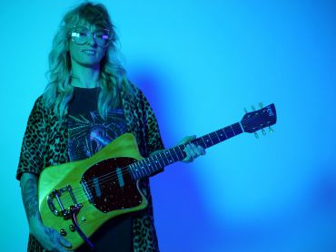 Woman with glasses playing an electric guitar under blue lighting.