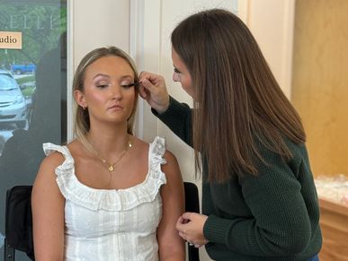 A makeup artist applies mascara to a seated woman in a studio setting.