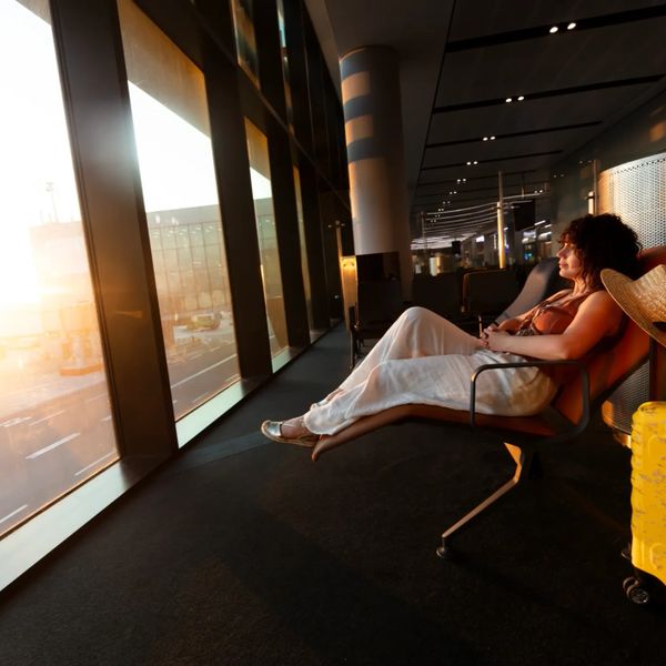Woman resting by airport window with yellow suitcase at sunset.
