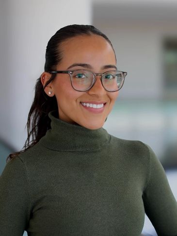 Young woman with glasses smiling, wearing a green turtleneck sweater.
