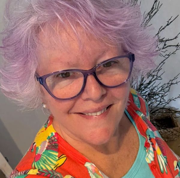Smiling woman with purple hair and glasses.
