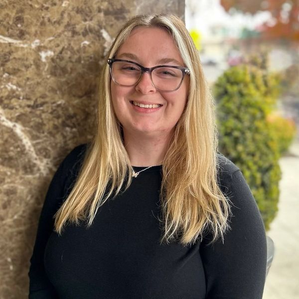 Smiling woman with glasses and long blonde hair stands against a marble wall.
