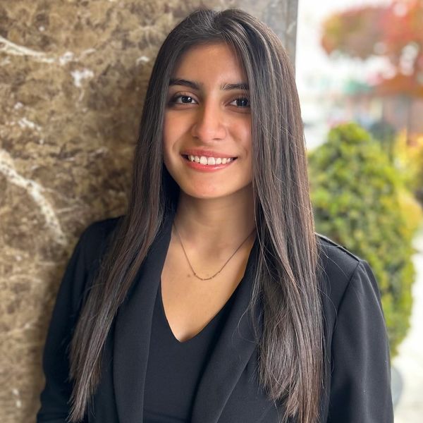 A young woman in black blazer smiling against a marble wall.