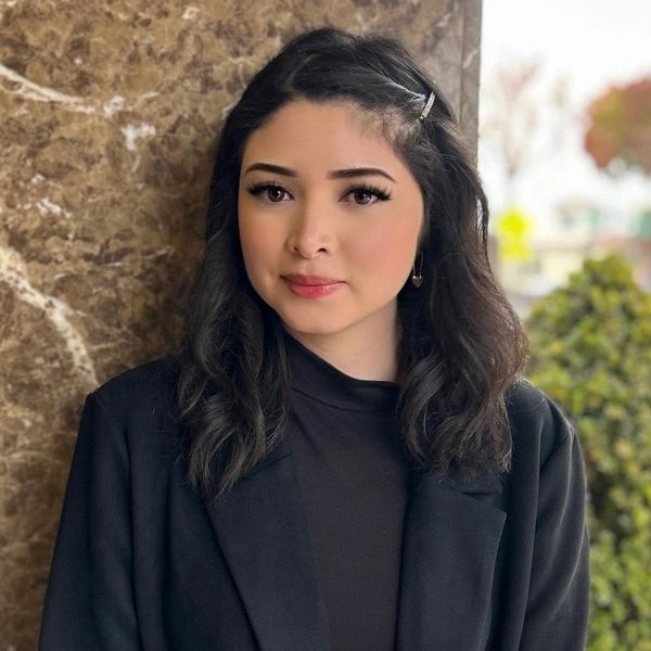 A young woman in a black coat stands against a marble wall outdoors.