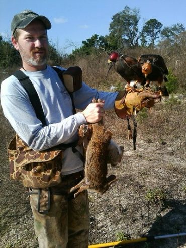 Man holding a dead rabbit and a falcon on his gloved hand outdoors.