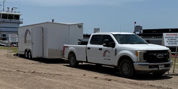 White pickup truck with trailer parked near a dock with warning signs about dangerous cargo.