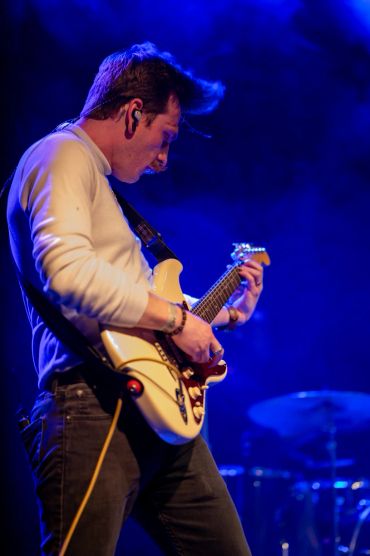 Man playing electric guitar on stage with blue lighting.