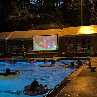 People watch a movie on a screen while floating in a pool at night.