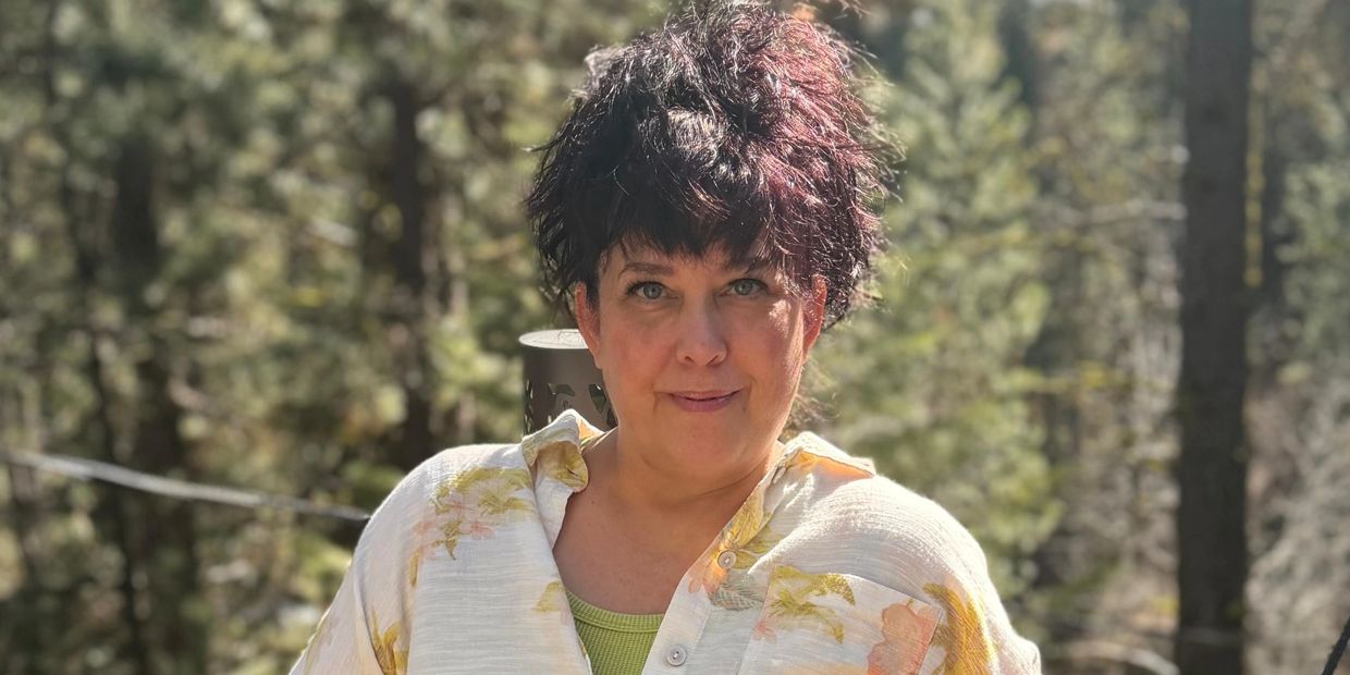 Woman with dark hair and floral shirt posing outdoors in sunlight.