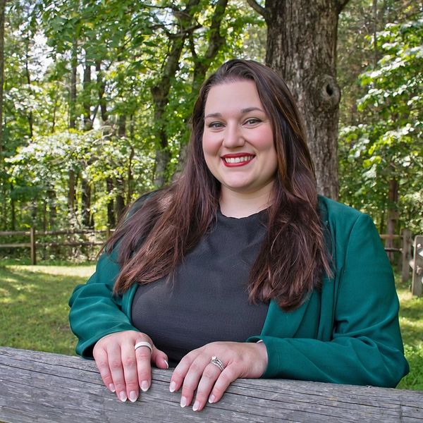 Smiling woman leaning on wooden fence in a green outdoor setting.