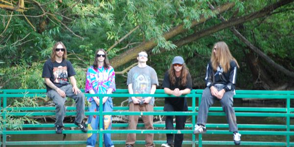 Five young adults with long hair and sunglasses on a green bridge in a wooded area.