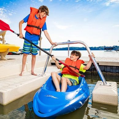 Two kids launching a kayak wearing life jackets standing on a dock surrounded by water.