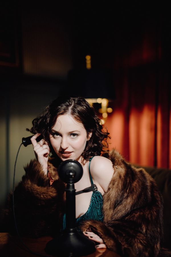 A woman in a fur coat speaks into a vintage telephone with an intense look.