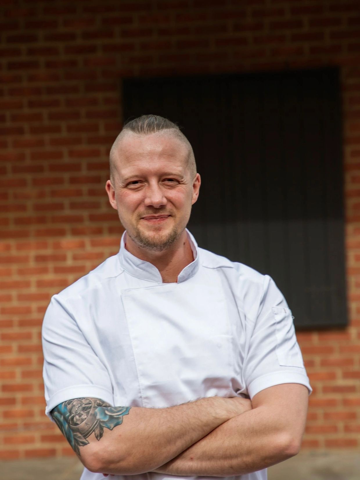 Smiling chef with tattooed arm in white uniform stands confidently.