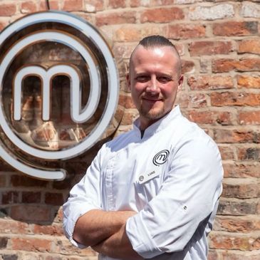 Chef posing confidently in front of MasterChef logo on brick wall.
