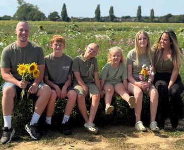 Family of six sitting outdoors with sunflowers, smiling happily.