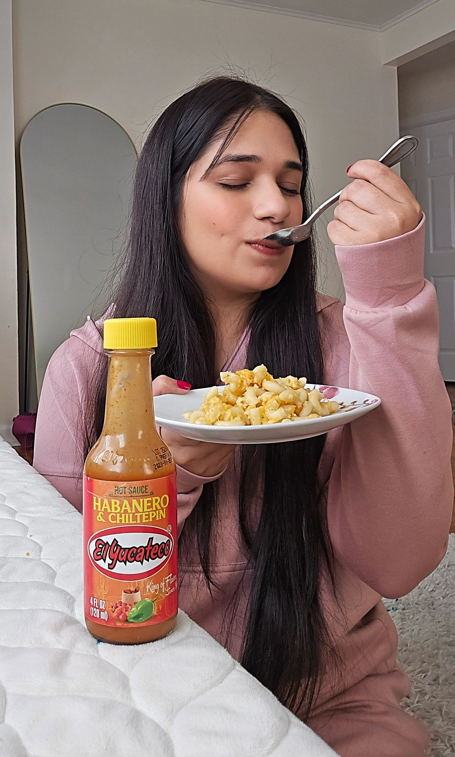 Woman enjoying macaroni with El Yucateco hot sauce in cozy setting.