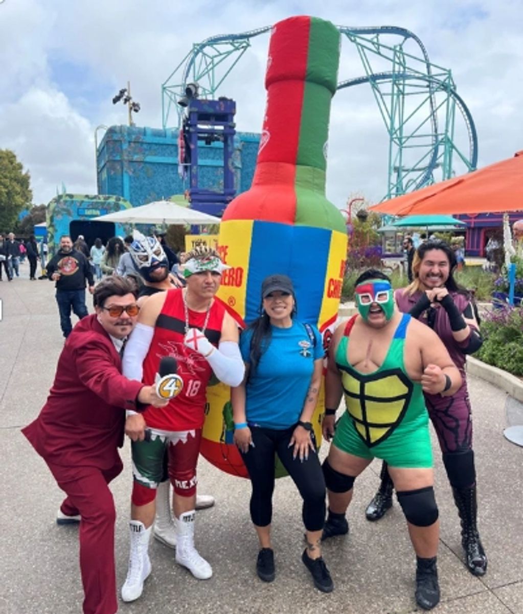 Group of people posing in colorful costumes at an amusement park with a large inflatable hot sauce bottle.