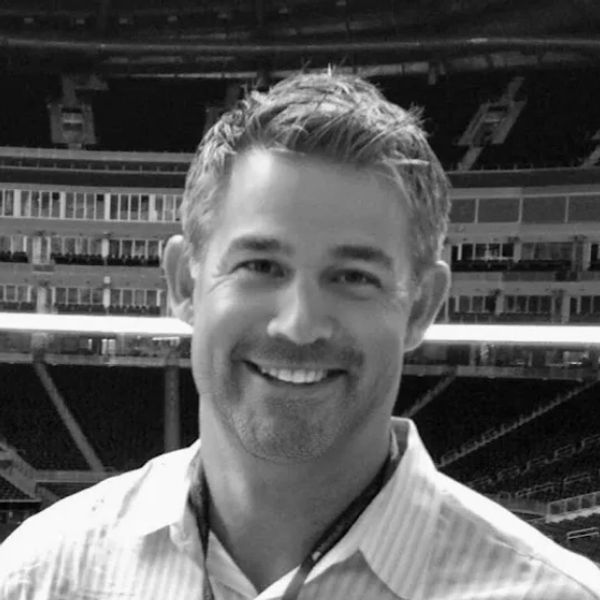 Smiling man in a stadium, wearing a light-colored shirt.