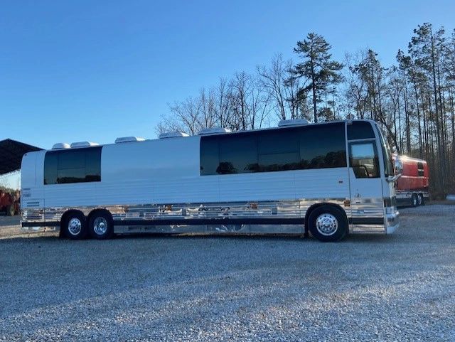 A sleek silver tour bus parked on gravel with trees in the background.
