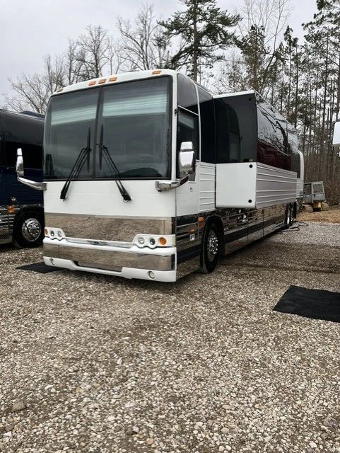 A large white and silver bus parked on a gravel lot with trees in the background.